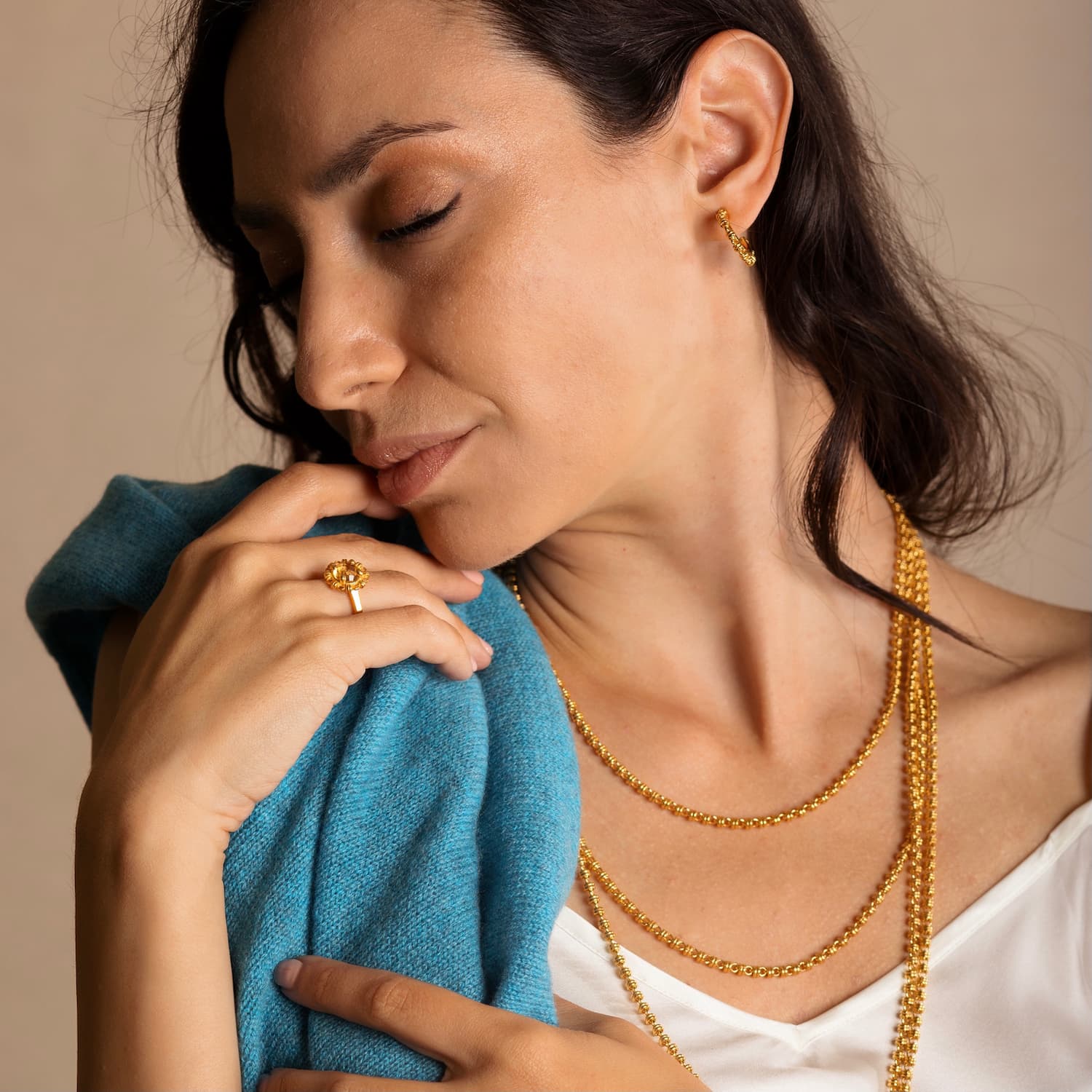 A model holding a turquoise sweater while wearing four gold chains in varying lengths from a short, 16-inch gold chain to a 36-inch gold chain with matching gold earrings and a gold ring in the same iconic chain design around a semi-precious gemstone. All hand-crafted Italian jewelry is made by DelBrenna Italian Jewelers in Tuscany.