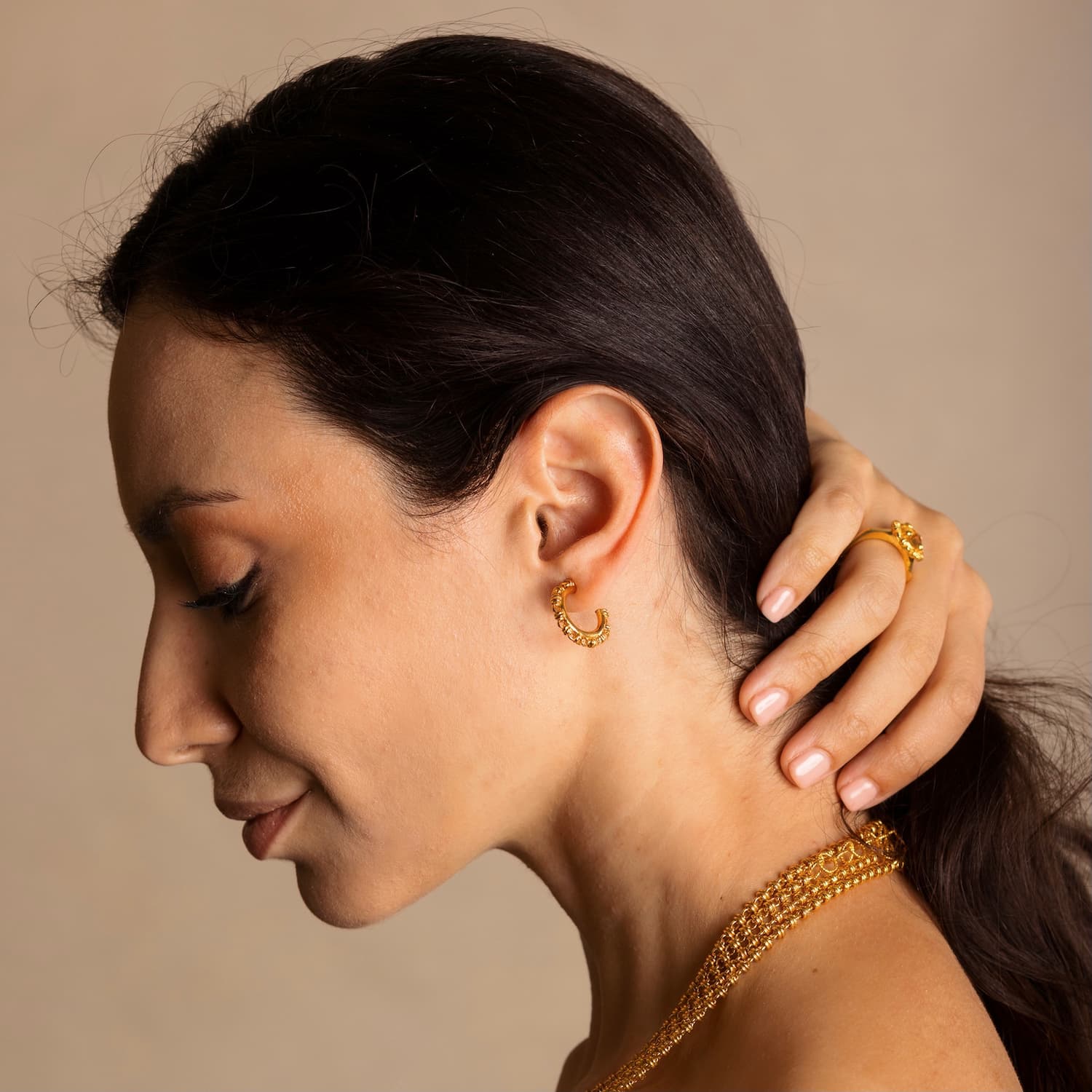 A model holding her hair back to show four gold chains with matching gold hoop earrings and a gold ring in the same iconic chain design around a semi-precious gemstone. All hand-crafted Italian jewelry is made by DelBrenna Italian Jewelers in Tuscany. 