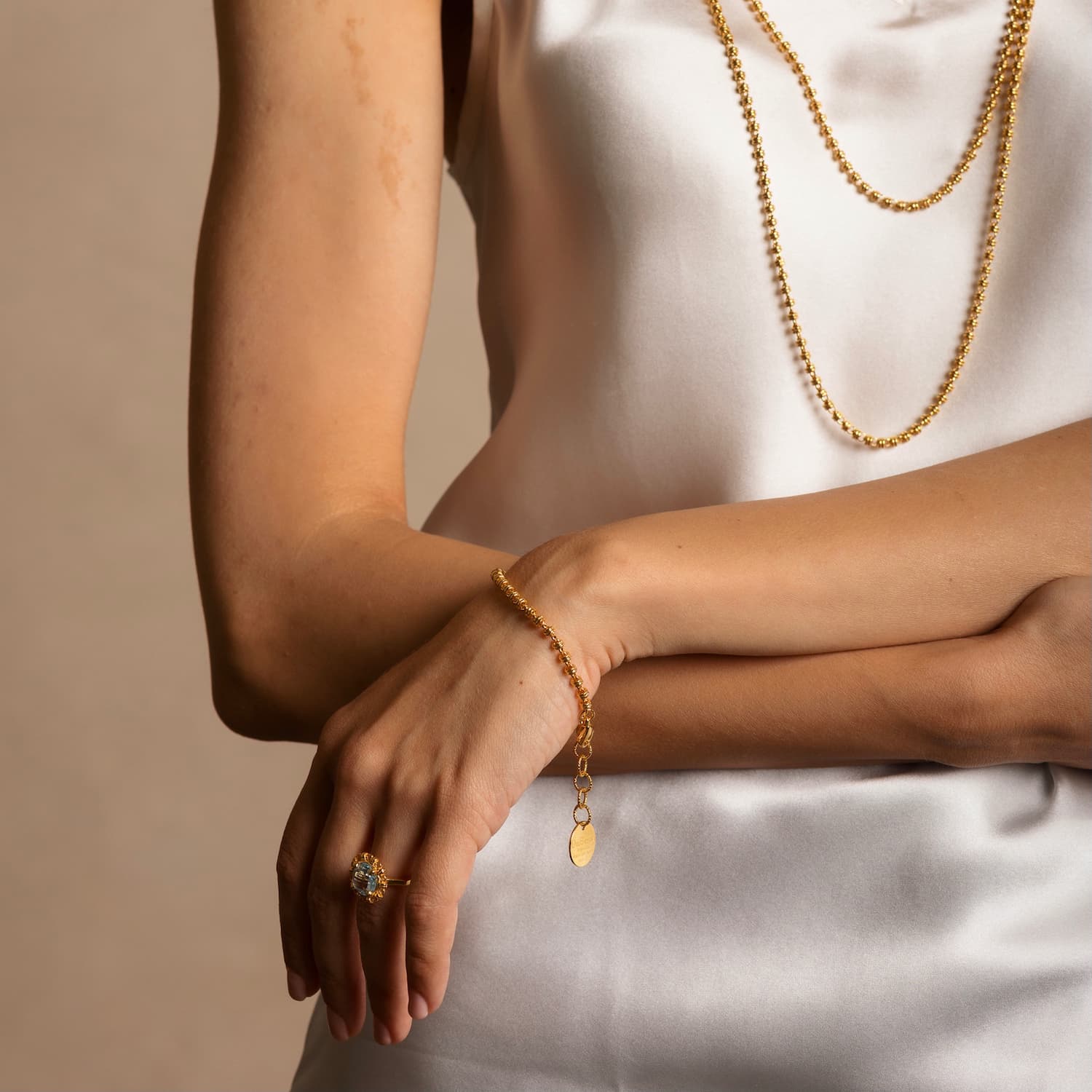 A model wearing a delicate gold chain bracelet with a small circular gold charm and lobster clasp all in 24K yellow gold finish with gold necklaces, earrings, and a ring to match. The jewelry is hand-crafted and designed from the iconic DelBrenna Links collection - Italian jewelry designs hand-crafted in Tuscany.