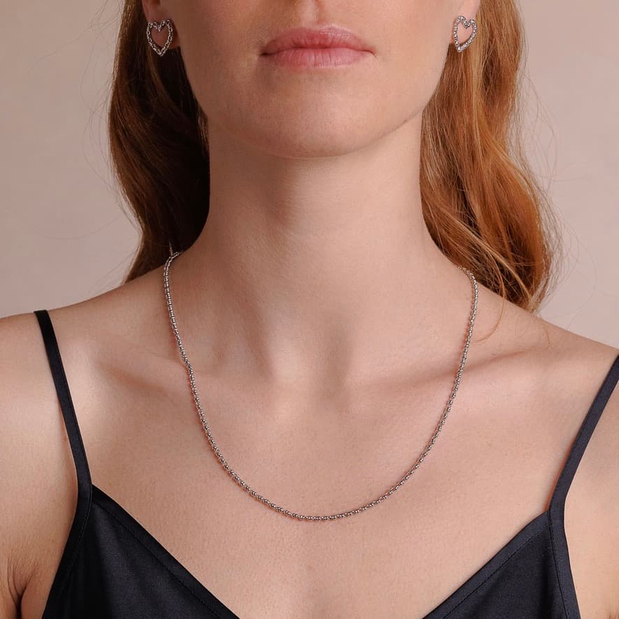 A front-facing, closeup view of a model wearing a short (18-inch) silver necklace with matching silver heart earrings. The necklace and earrings are a delicate 2MM version of the iconic silver chain designed by DelBrenna Italian jewelry designers in 1974 in Tuscany.
