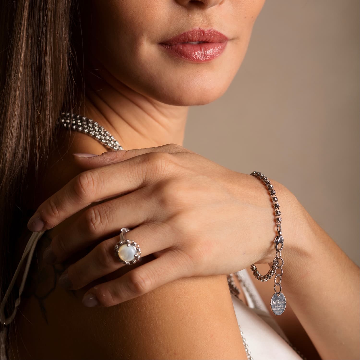 A close-up view of a model wearing a stunning silver bracelet in a silver chain design with a silver charm with the DelBrenna name - along with a matching silver chain necklace and a ring with the silver chain designed around a gemstone. All jewelry is hand-crafted by DelBrenna Italian Jewelry designers in Tuscany. 