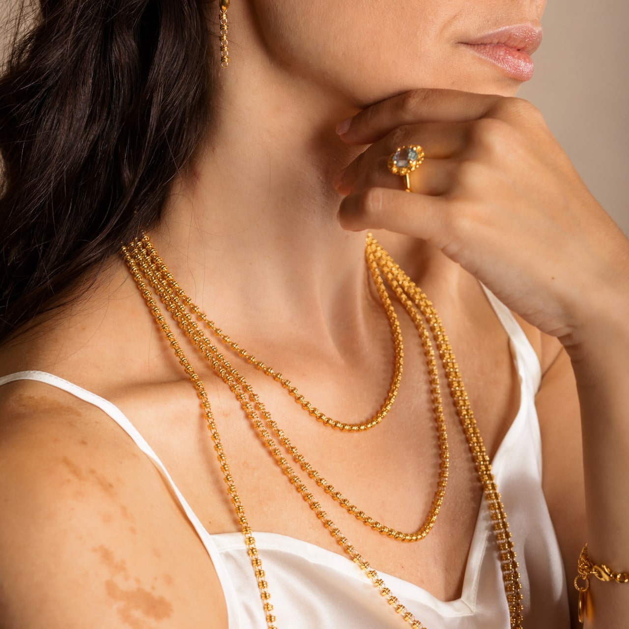 A person modeling four gold chains in varying lengths from a short, 16-inch gold chain to a 36-inch gold chain with matching gold earrings and a gold ring in the same iconic chain design around a semi-precious gemstone. All hand-crafted Italian jewelry is made by DelBrenna Italian Jewelers in Tuscany. 