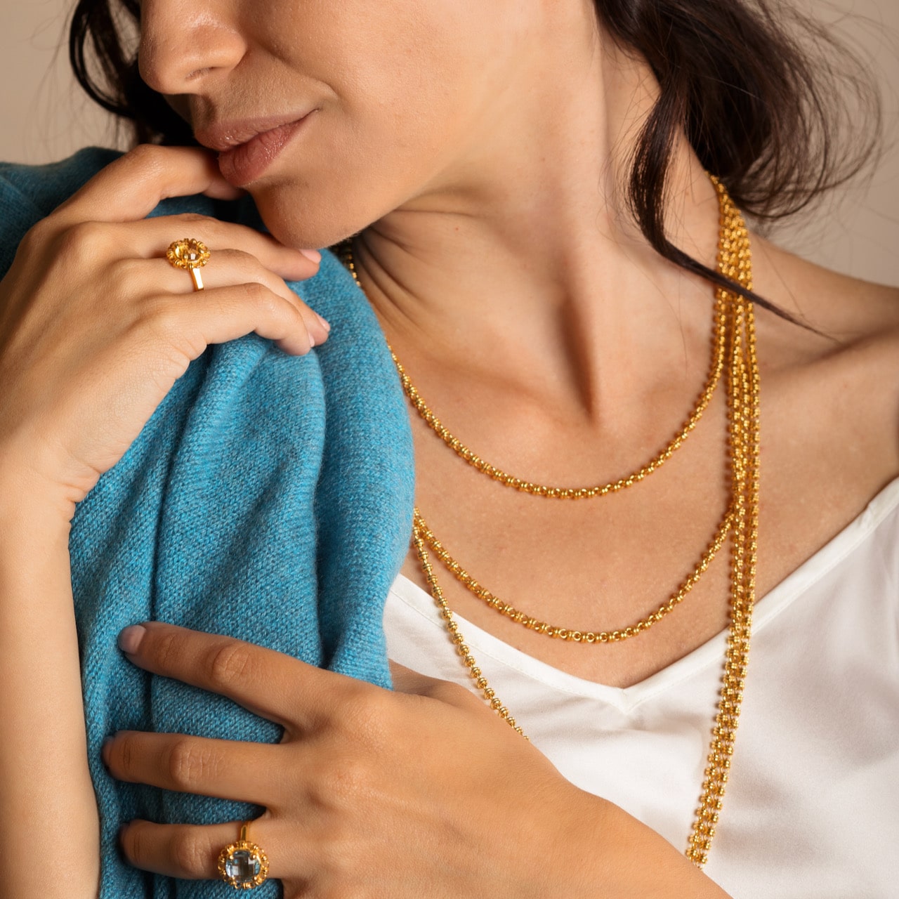 A model holding a turquoise sweater while wearing four gold chains in varying lengths from a short, 16-inch gold chain to a 36-inch gold chain with matching gold earrings and a gold ring in the same iconic chain design around a semi-precious gemstone. All hand-crafted Italian jewelry is made by DelBrenna Italian Jewelers in Tuscany. 