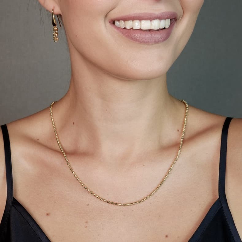 A front-facing, closeup view of a model wearing a short (18-inch) gold necklace with matching gold earrings. The necklace and earrings are a delicate 2MM version of the iconic gold chain designed by DelBrenna Italian jewelry designers in 1974 in Tuscany. 