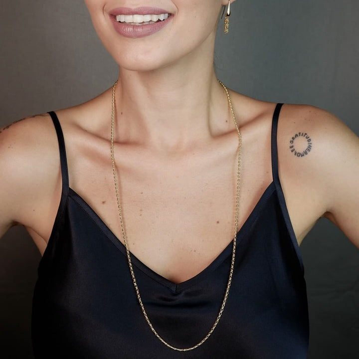 A front-facing, closeup view of a model wearing a long (20-inch) gold necklace with matching gold earrings. The necklace and earrings are a delicate 2MM version of the iconic gold chain designed by DelBrenna Italian jewelry designers in 1974 in Tuscany. 