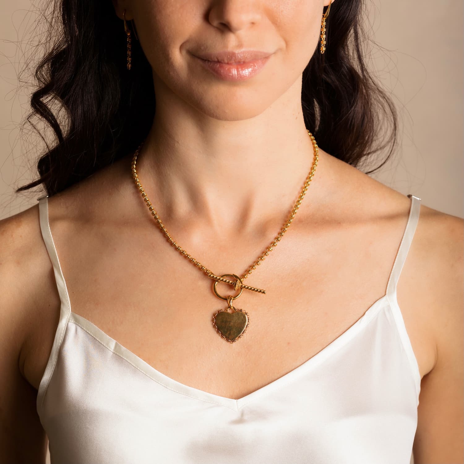 A model wearing a gold necklace with a toggle clasp with a gold heart pendant attached and gold earrings to match the gold chain in the necklace design - all are from the Links 3mm collection from DelBrenna Italian Jewelry designers and artisans. Hand-crafted in Tuscany.