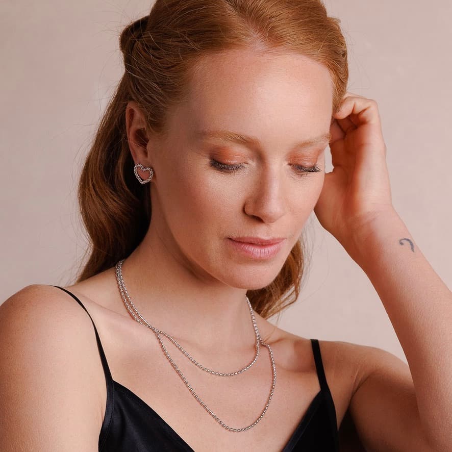 A model wearing two delicate, hand-crafted 2MM silver chains in two different lengths: a short, 16-18” silver chain necklace and a 20-22” silver necklace with matching silver heart earrings and the Links 2MM silver bracelet with a silver charm  - all jewelry is crafted by DelBrenna Italian Jewelry designers in Tuscany.